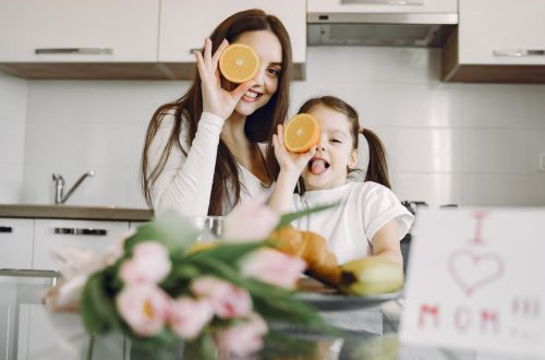 From below of cheerful mother and daughter in domestic clothes smiling and playing with oranges while sitting together at table with bouquet of tulip and drawing of child